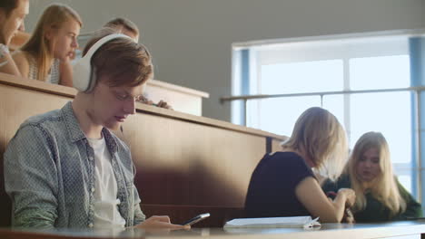 A-male-student-in-a-University-auditorium-listens-to-music-in-white-headphones-during-a-break-without-paying-attention-to-others.-A-lot-of-people-in-the-audience-are-talking.