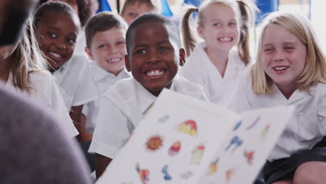 male teacher reading story to group of elementary pupils wearing uniform in school classroom