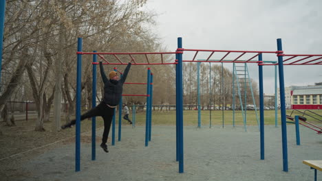 a coach leaps up to grab a red iron bar and steadily moves across it, after reaching the end of the bar, he drops down, in the background, a boy is playing football in a stadium
