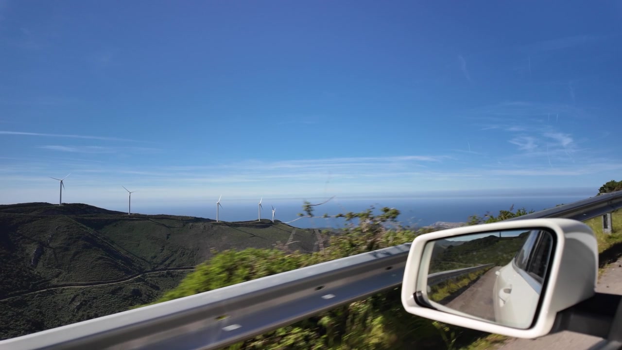Car window view of wind turbines on green mountains above the sea