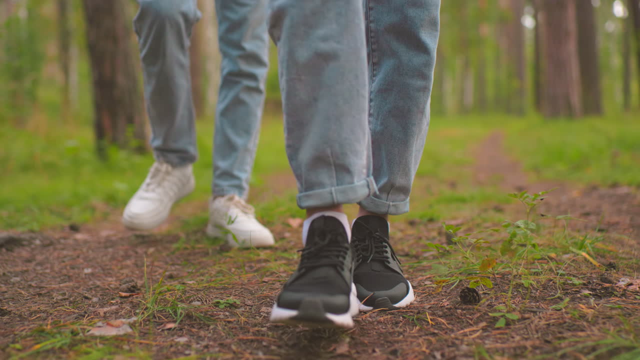 Close-up of two ladies walking along a forest trail, each wearing blue jeans, one in white sneakers and the other in black, their steps move in sync over a path covered with pine cones and leaves