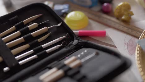 A set of clay sculpting tools are nestled in their container, on a table that has various toys and crafting objects.