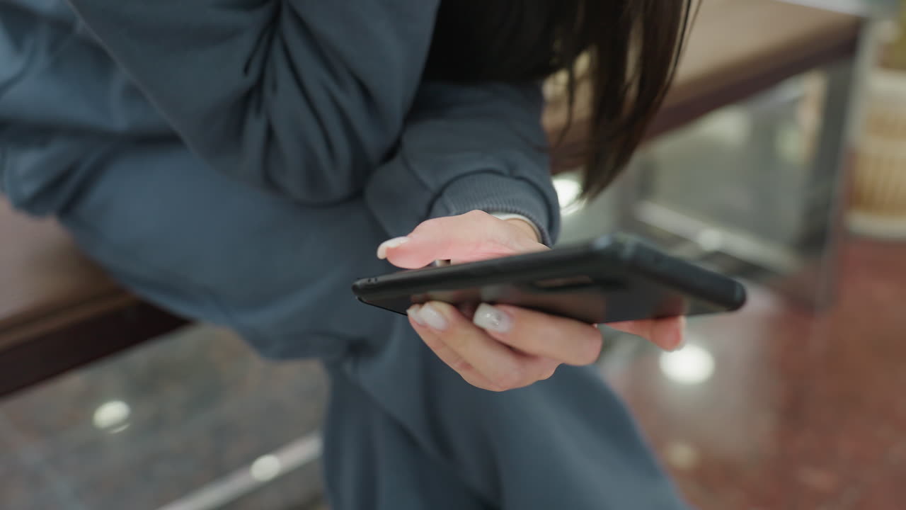 Close up of young woman in dark casual tracksuit sitting indoors, holding smartphone with both hands, leaning slightly forward while browsing or texting, soft indoor light and background blur visible