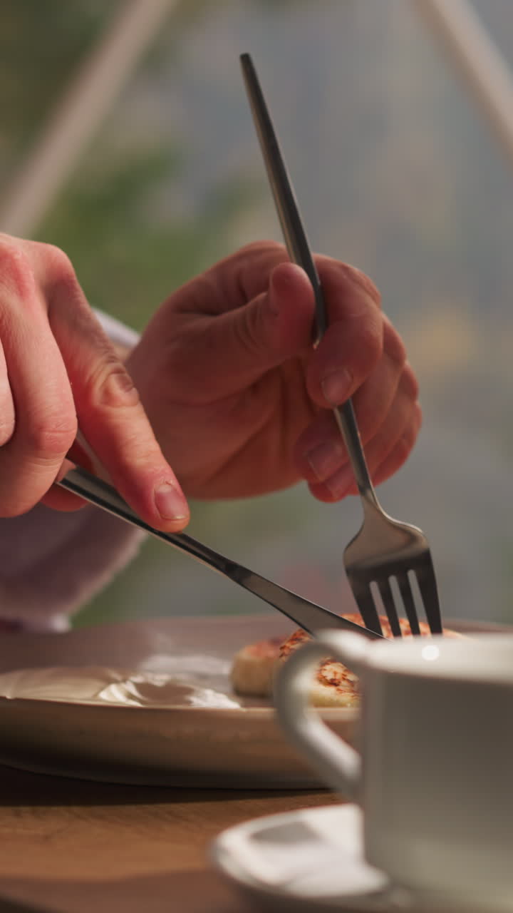 Man cuts piece of pancake at table in glamping house. Young man eats sweet breakfast against glass wall in eco-hotel. Standard room service breakfast