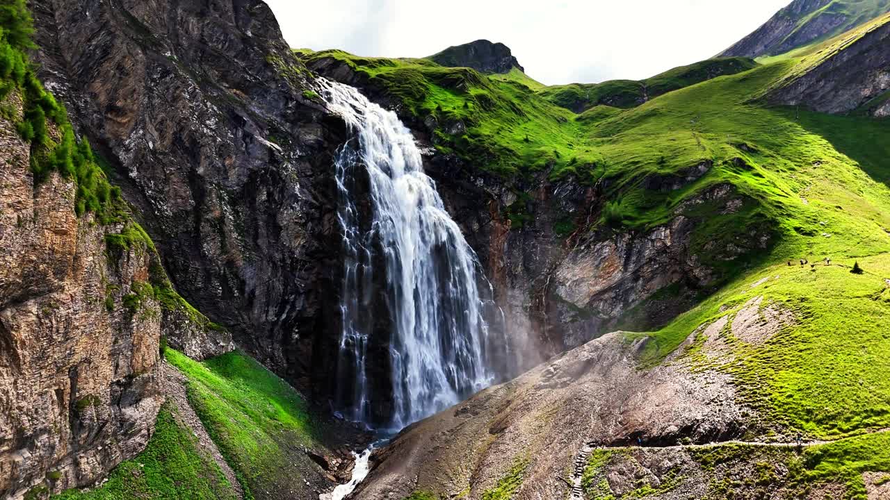 In Adelboden, Switzerland, the Engstligenfälle waterfall drops from towering alpine cliffs into a valley of green slopes, where rugged peaks, forests, and snowy ridgelines form a mountain scene