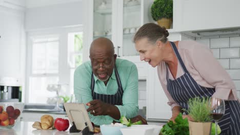 smiling senior diverse couple wearing blue aprons and using tablet in kitchen