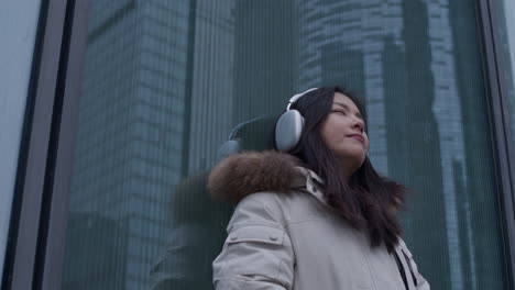 young asian girl listens to music in headphones outdoors, leaning against building with skyscraper reflection in window