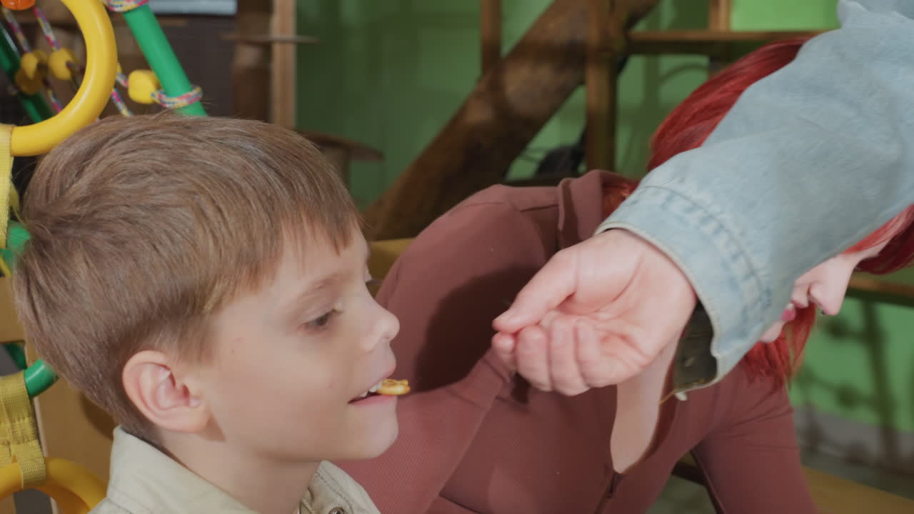 Un ragazzo caregiver in una stanza dei giochi riceve uno snack mentre una logopedista dai capelli rossi incoraggia l'uso della parola; anelli tattili e attrezzature con corde visibili; primo piano di un bambino attento, sorridente e concentrato; atmosfera calorosa e di supporto