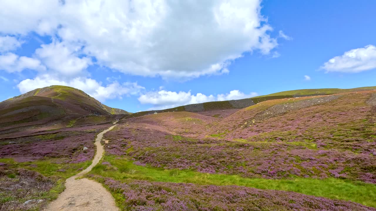 A steady camera moves forward along a scenic mountain trail in Glen Clova, revealing rolling hills, heather, and dramatic skies under bright daylight
