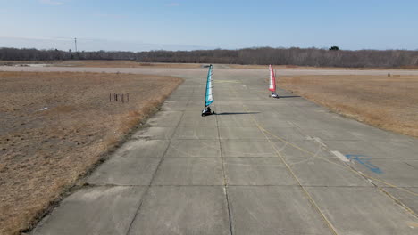 moving backward drone footage tracking two weaving land sailing buggies on disused airfield