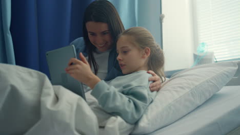 Smiling-mom-visiting-daughter-play-on-tablet-computer-together-in-hospital-room