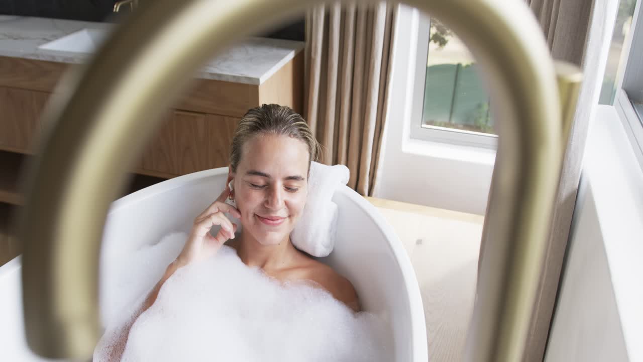 Adult woman receiving call, holding smartphone then switching to earbuds while relaxing in oval tub