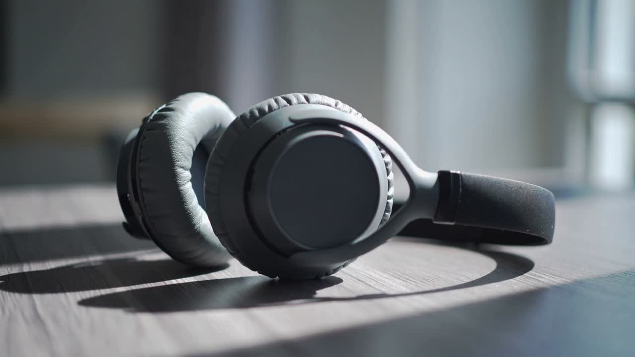 Kameraet trekker seg tilbake og fokuserer på matte over-ear hodetelefoner på et trebord, og avslører lys fra et vindu.