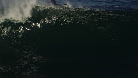 telephoto tracking pan across huge ocean wave as surfers in full wet suits paddle over top away, slow motion