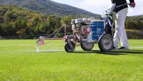 máquina de pintura de línea siendo empujada por el operador para pintar líneas en el campo de fútbol con montañas en el fondo