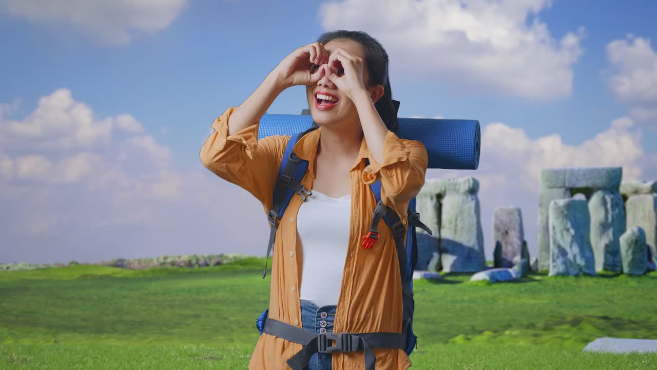 Asian Female Hiker With Mountaineering Backpack Smiling And Making Binoculars Gesture Then Looking Around While Traveling In Stonehenge