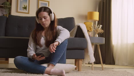 woman wearing wireless headphones sitting on floor at home streaming music from mobile phone 1