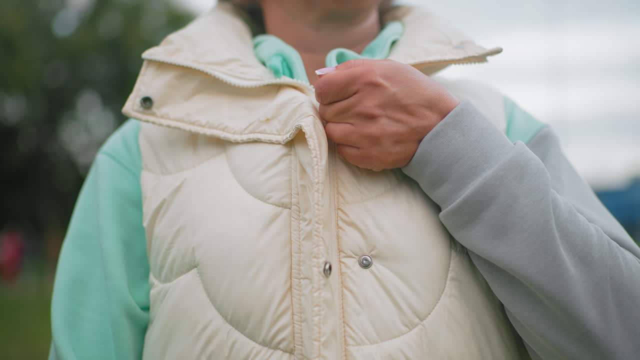 Close up of animal instructor closing light puffy jacket over mint green hoodie outdoors on grassy field, adjusting clothing for warmth on cool cloudy day, preparing for outdoor pet training session
