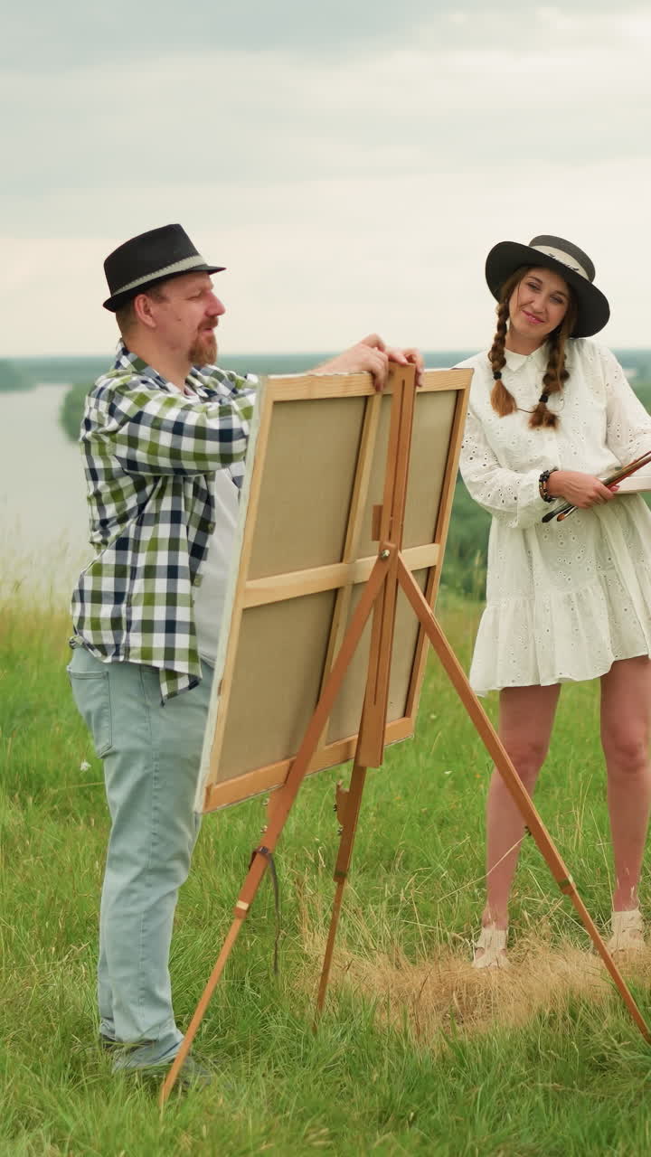 두 파트너가 잔디에서 보입니다. 검은 모자와 카레드 셔츠를 입은 남자는 나무 삼각대 위에 그림판을 세우고 있습니다. 근처에  드레스를 입은 여자가 웃으며 서 있습니다.
