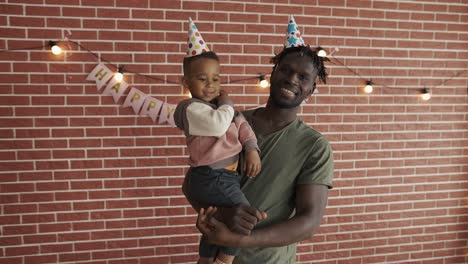 Portrait-of-cute-afro-american-boy-on-father's-arms-in-b-day-hats