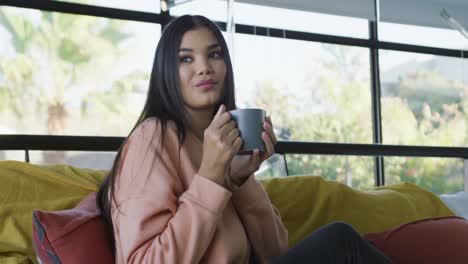 video of happy biracial woman sitting on sofa and drinking coffee