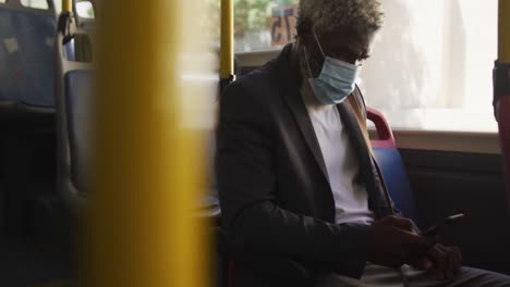 Afroamerikanischer-älterer-Mann-Mit-Gesichtsmaske-Und-Smartphone,-Während-Er-Im-Bus-Sitzt