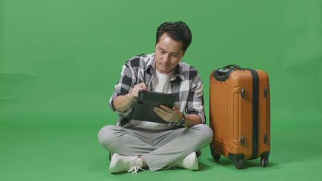 full body of asian male traveler with luggage using a tablet while sitting in the green screen background studio