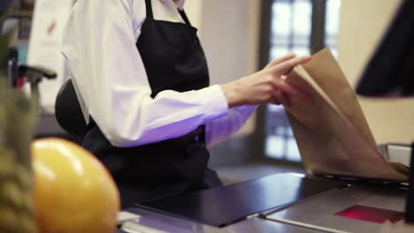 Unrecognizable-Slender-Saleswoman-In-White-Shirt-And-Black-Apron-Scanning-Product,-Fruits-At-Checkout-Counter-In-Store-And-Putting-It-Into-Brown-Paper-Bag