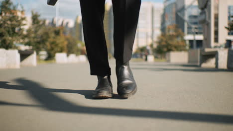 leg view of individual wearing black trousers and shoes walking confidently outdoors on tarred road, shadow cast on ground, surrounded by trees and urban background