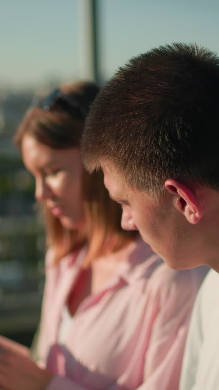 primer plano de una mujer joven mostrando un libro a su amigo masculino sentado al aire libre, su atención desviada a otra dirección, fondo borroso con vegetación y paisaje urbano