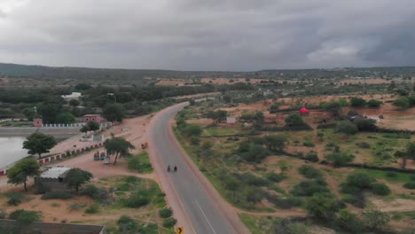 Die-Drohne-Nimmt-Zwei-Radfahrer-Auf,-Die-Ihr-Fahrrad-Auf-Der-Abgelegenen-Autobahn-Von-Tharparkar-Fahren,-Die-Von-Sand,-Gras-Und-Sträuchern-Umgeben-Ist