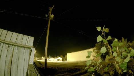 Lapso-De-Noche---Mirando-El-Cielo-Desde-Un-Campo-Con-Un-Edificio-Antiguo-Y-Una-Valla-De-Madera-En-Un-Pequeño-Pueblo