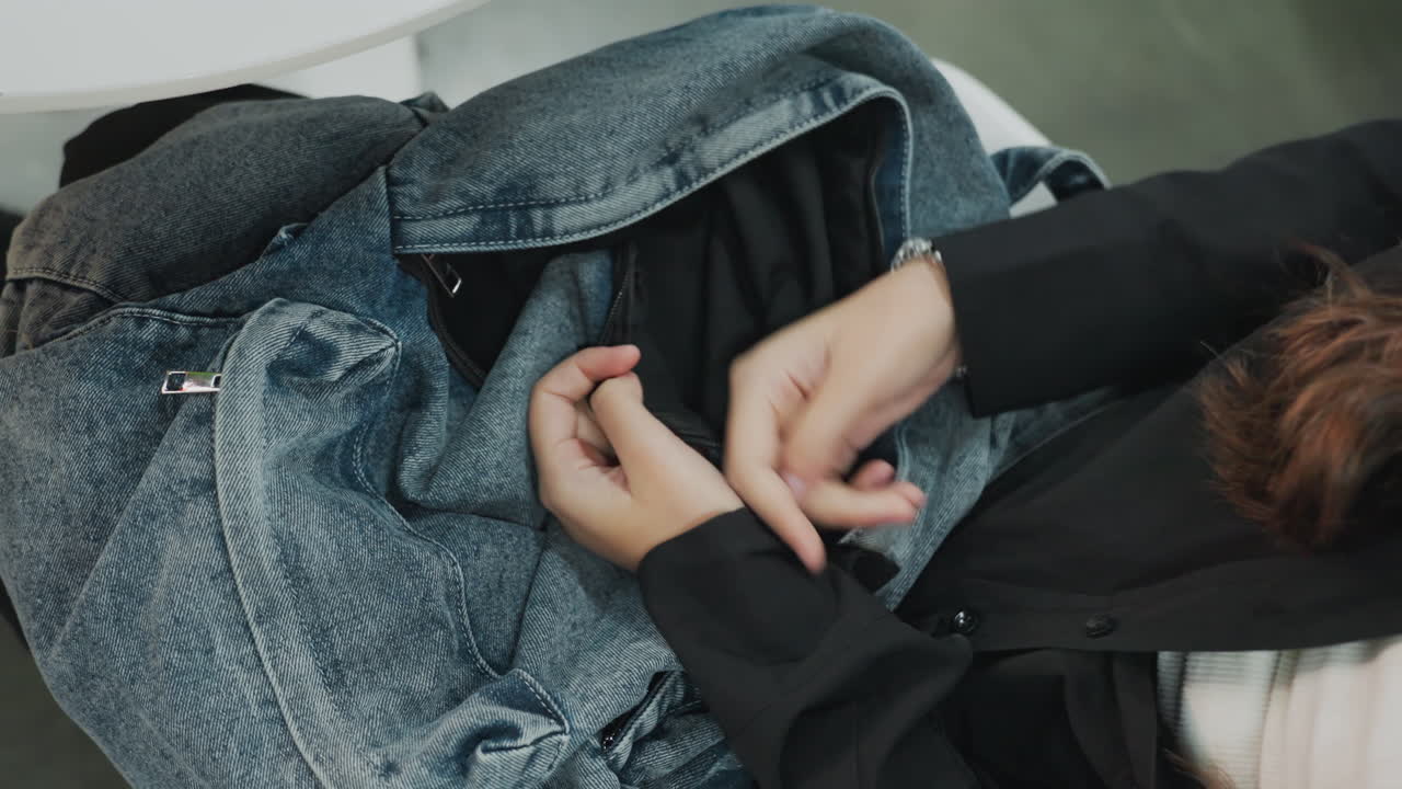 overhead view student hands searching backpack, denim bag with teal case visible, zipper and pocket accessed while seated in campus waiting area, fingers fidgeting with bracelet as items are checked