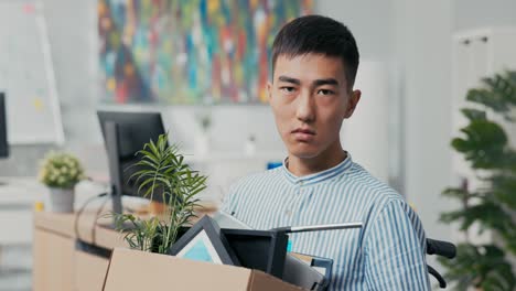 disabled ambitious man in shirt with asian korean beauty sits in wheelchair at desk in the company on knees holding cardboard box with packed things, gets fired from job unable to fulfill his duties