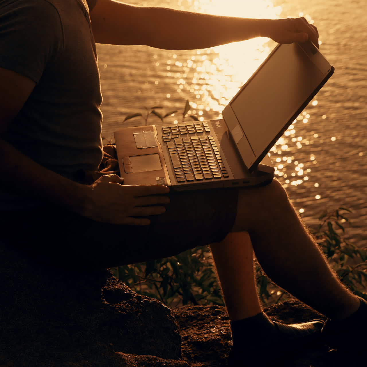 A man with a laptop in his hands is sitting on a cliff the background of the reflection of the sun path along the river at sunset.