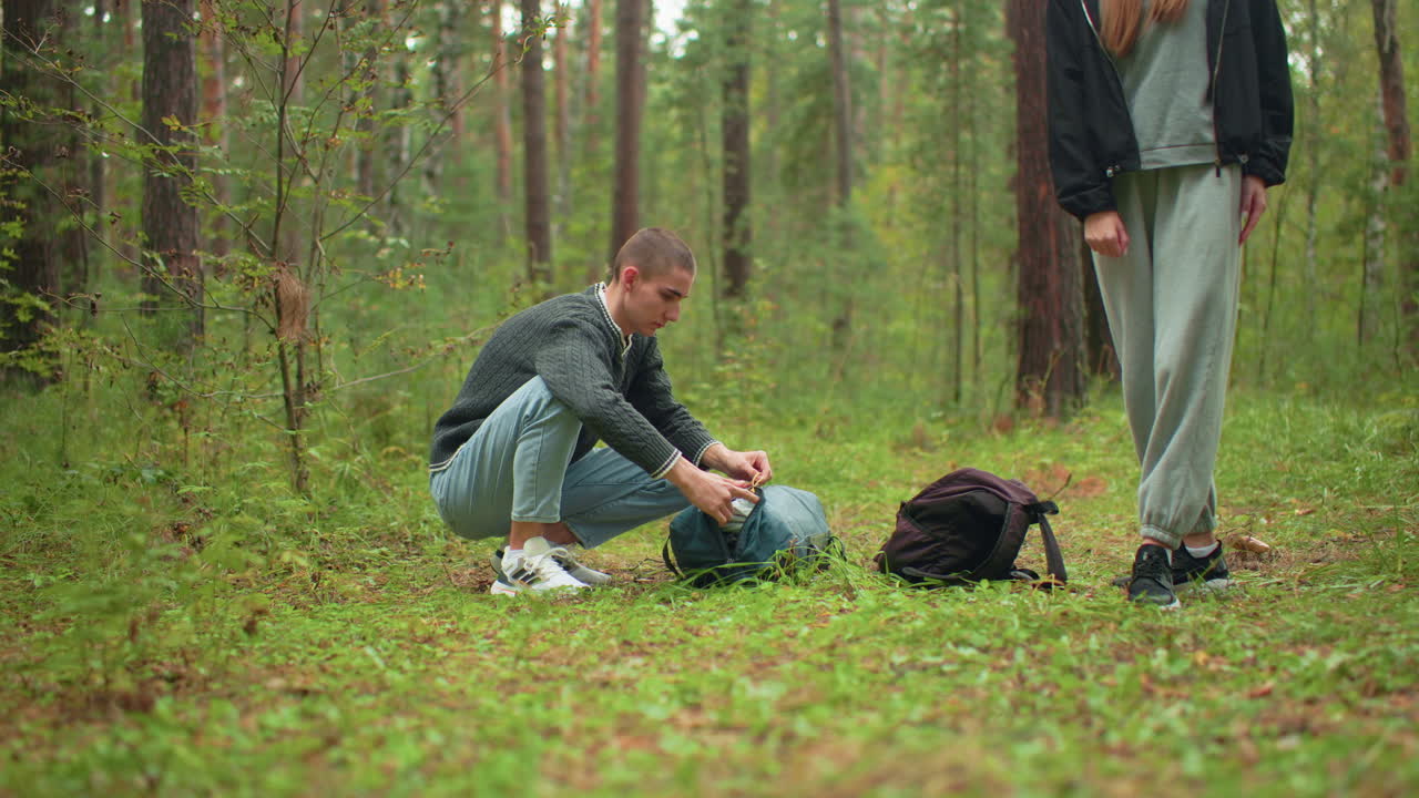 boy crouched in green forest opening bag to unpack tent as woman in tracksuit stands nearby stretching arms, surrounded by trees and grass during calm outdoor moment in woodland setting