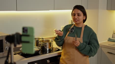 a happy brunette girl in a green jacket and a beige apron runs her culinary blog and uses her phone to tell how to properly prepare food and other culinary secrets in a modern kitchen