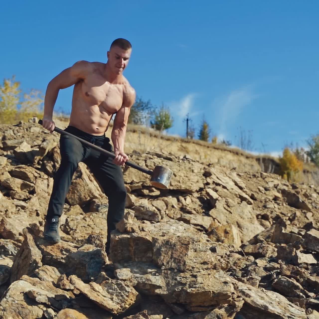 Muscular bodybuilder is training with steel hammer on hill. Strong athlete without shirt breaking stones by a metal tool outdoors. Slow motion.