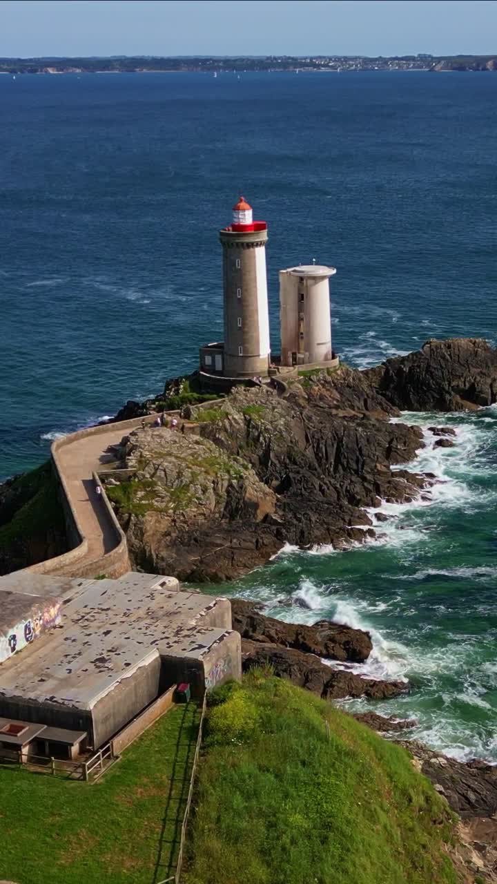 Petit Minou Lighthouse on rocky islet, stone tower, causeway, and blue sea with waves, Plouzané, Brittany, France. Aerial forward, vertical format