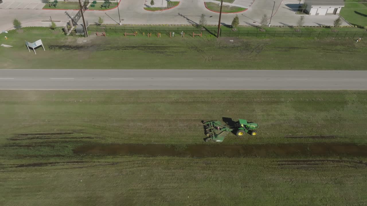 A 4K aerial drone view captures a heavy equipment operator driving a tractor with a brush hog mower attachment, mowing the median of Fairmont Parkway on a sunny morning in La Porte, Texas.