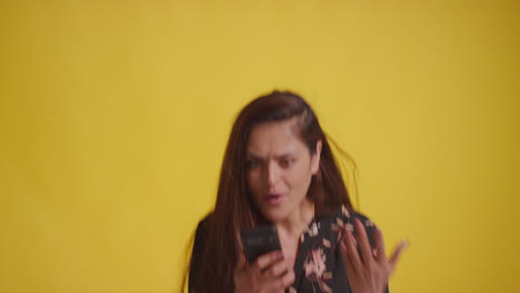 studio shot of excited woman with mobile phone celebrating winning money against yellow background