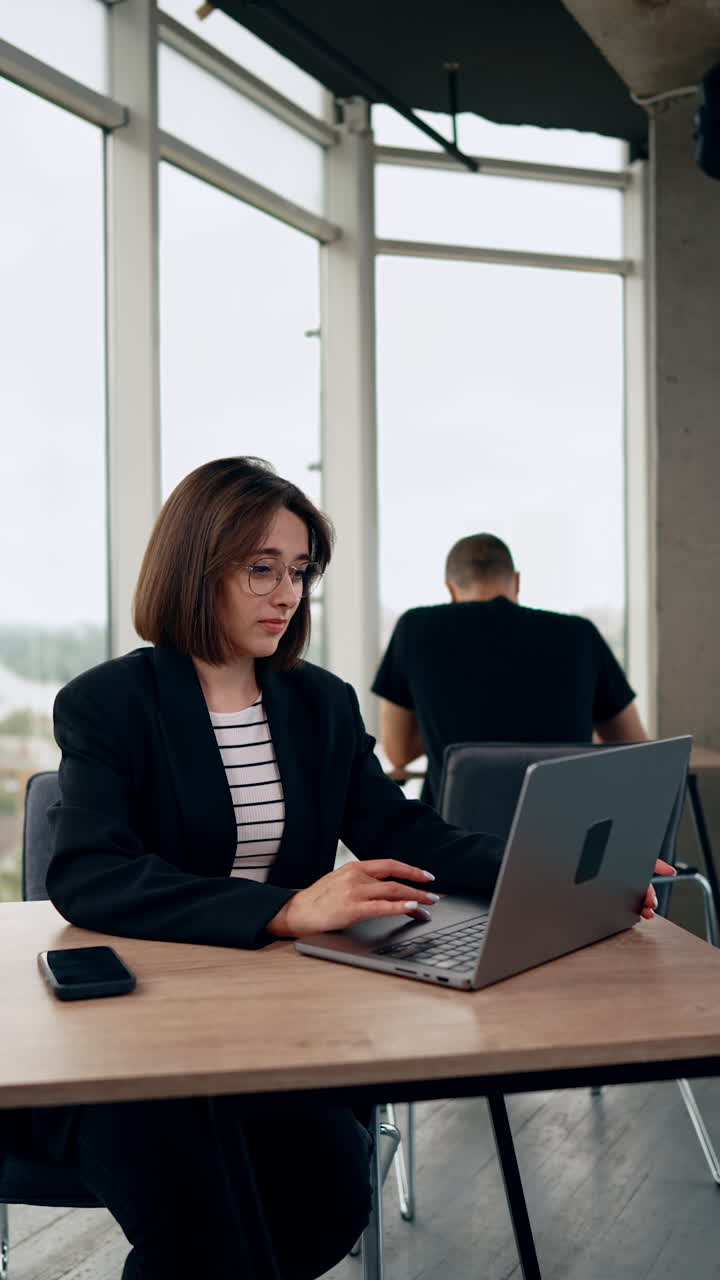 Brunette woman sitting at desk opens her laptop. Business lady starts work on laptop. Man sits at backdrop. Vertical video.