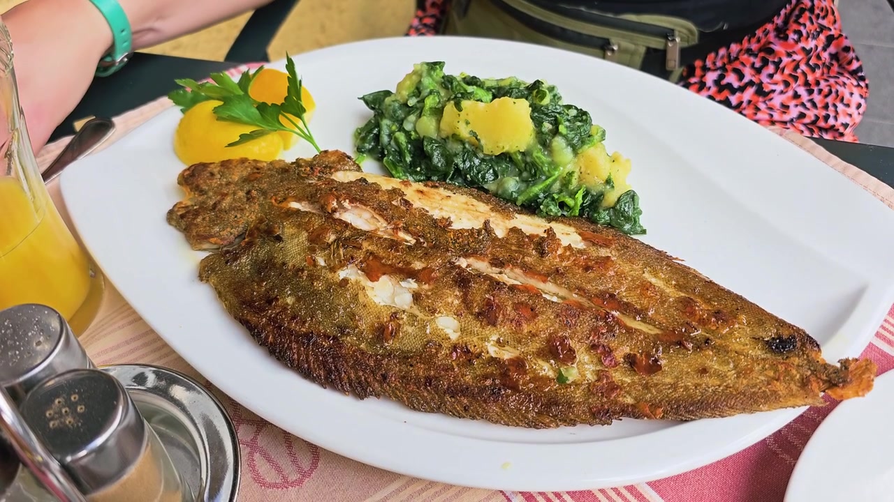 Frischer, in der Pfanne gebratener Seezungenfisch, ganz in einem Restaurant serviert, mit knuspriger, gewürzter Haut, Spinat-Kartoffel-Beilage, Zitronengarnitur, Petersilie und Getränken auf einem sonnigen Terrassentisch, ein Gourmet-Meeresfrüchte-Mahl.