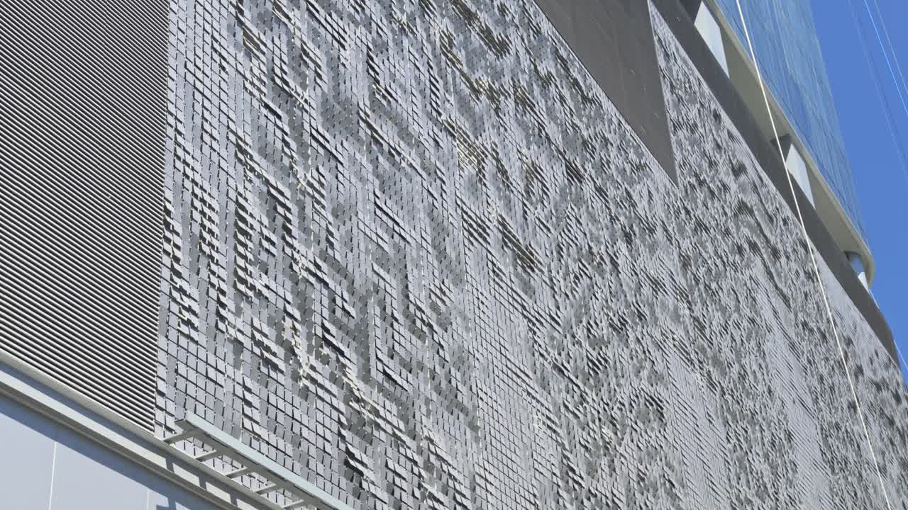 A textured surface resembling waves or ripples, part of a building situated in Brickell, Miami, glimmers under a clear, sunny sky, adding a modern architectural touch to the vibrant area.