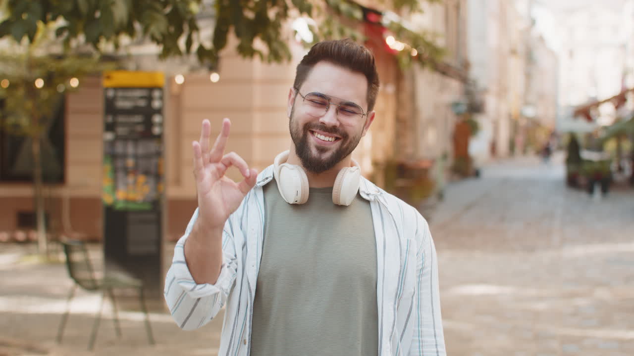 retrato de un joven millennial guapo mirando a la cámara mostrando la mano de un letrero ok en la calle de la ciudad