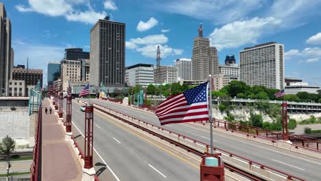 saint paul, el horizonte de minnesota en un día soleado, mostrando edificios altos, un puente con banderas estadounidenses, y una orilla del río bajo un cielo azul claro