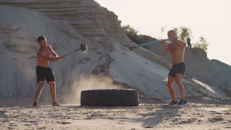 Zwei-Männliche-Athleten,-Die-Gemeinsam-Trainieren,-Schlagen-Bei-Sonnenuntergang-In-Den-Bergen-Mit-Einem-Hammer-Auf-Den-Sand.-Ausdauertraining