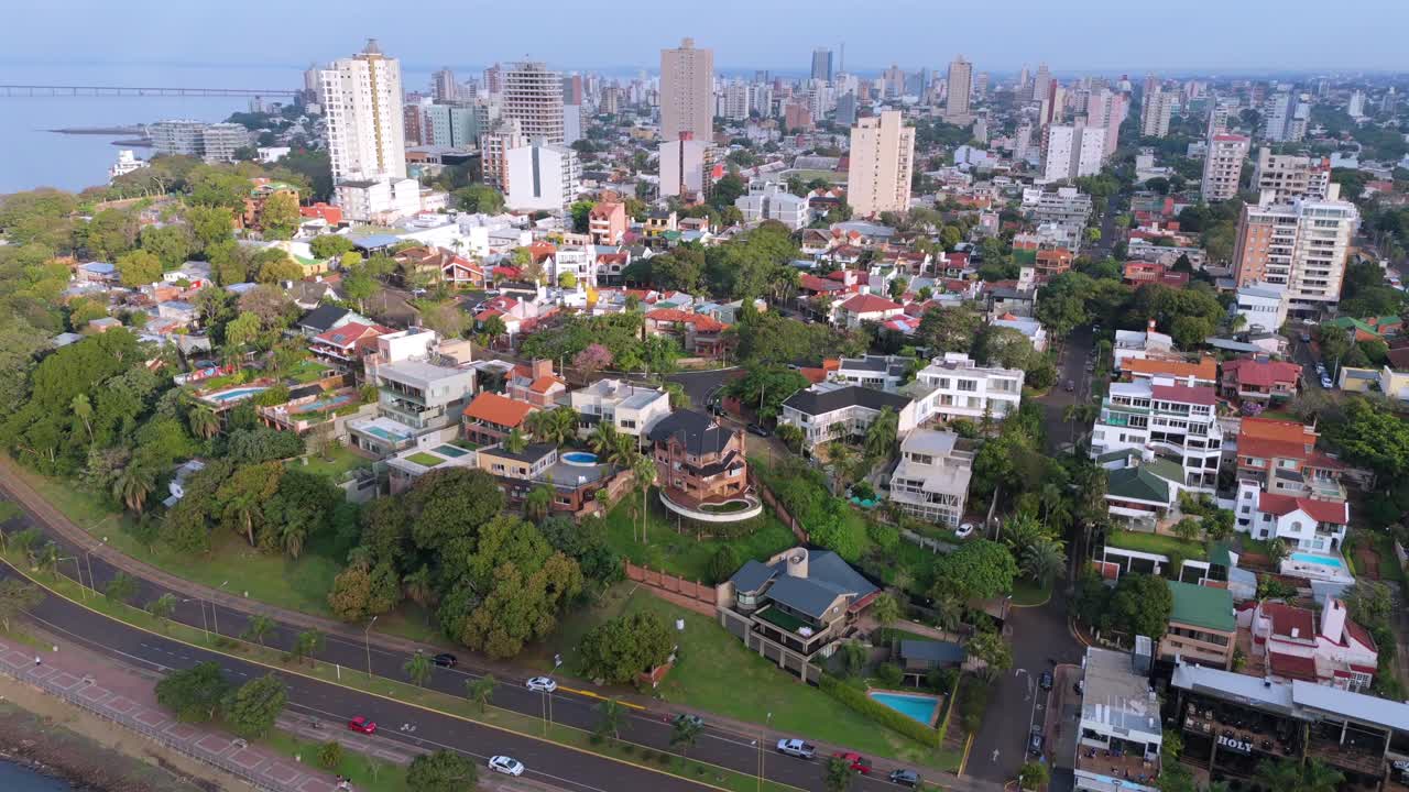 Vista aerea da drone di una zona residenziale con case moderne, vegetazione lussureggiante e skyline di grattacieli di Posadas, capitale della provincia di Misiones in Argentina, sotto la soffice luce del sole mattutino