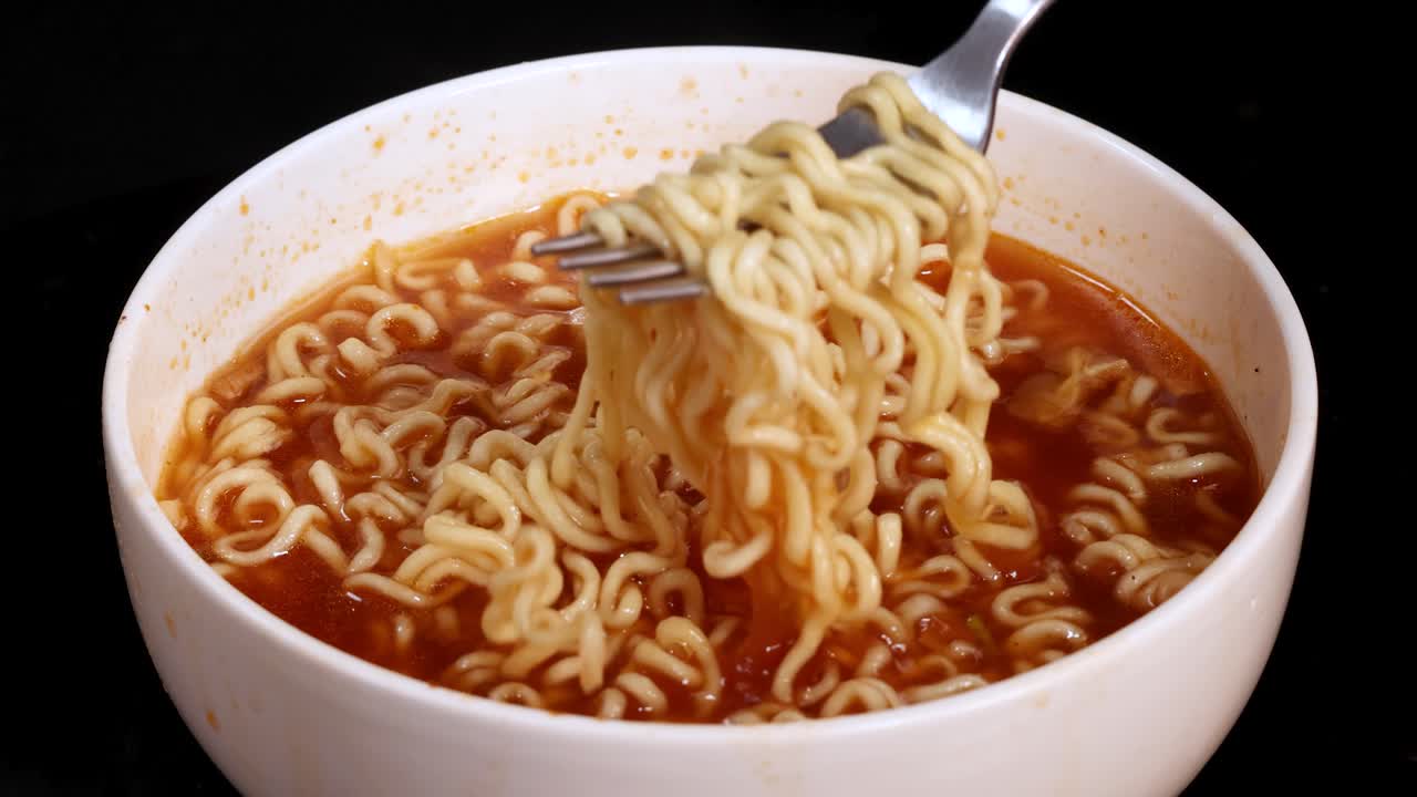 A hand uses a fork to lift and twirl cooked instant noodles from a steaming bowl of broth, under bright studio lighting with a black background
