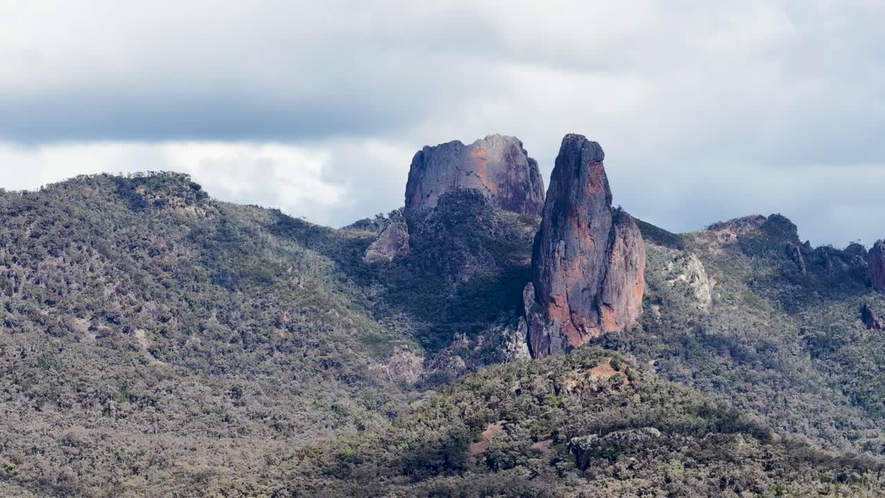 Static wide shot of the Breadknife rock spire and surrounding forested mountains under cloudy daylight, with no visible camera movement or human presence